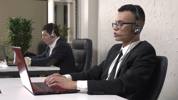 Young successful African businessman wearing glasses sits at the office and working on laptop, with headset, Caucasian man in suit in the background. Call center concept. 60 fps - Materiaali, video