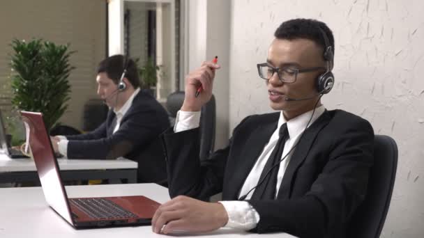 A young successful African man is sitting in the office and talking on the headset, call center concept. Caucasian man in suit in the background. Call center concept. 60 fps - Кадри, відео