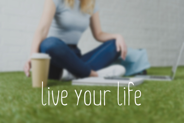 cropped shot of woman sitting on grass with cup of coffee to go and working with laptop, live your life inscription - 写真・画像