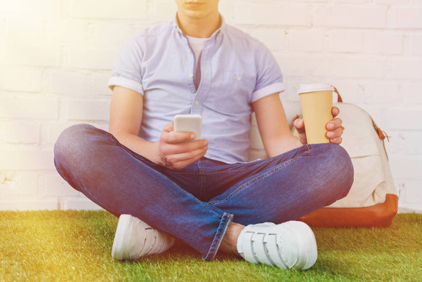 recortado disparo de joven sentado en la hierba con teléfono inteligente y café para ir
 - Foto, Imagen