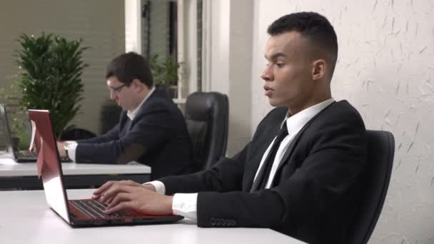 A young successful African male businessman working in the office on a laptop, tired, holding his head, a headache concept. Caucasian man in suit in the background. 60 fps - Imágenes, Vídeo