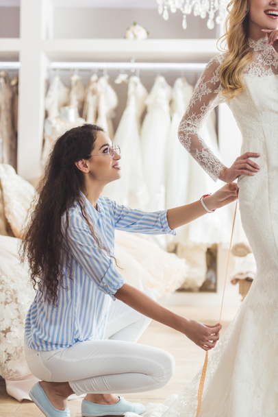 Beautiful bride and female tailor trying on dress in wedding salon - Foto, afbeelding