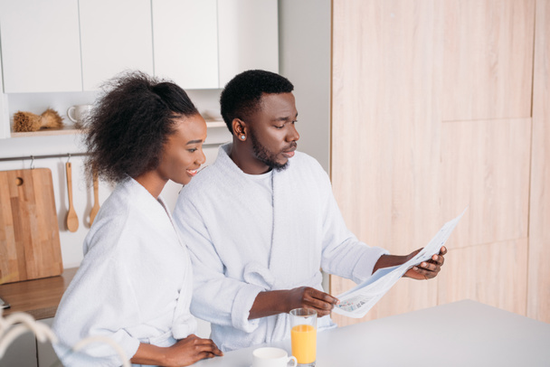 African american man reading newspaper and smiling girlfriend in kitchen - Foto, immagini