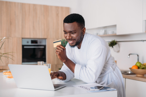 Usmívající se muž dělá online nakupování a stojí s kreditní kartou v kuchyni - Fotografie, Obrázek