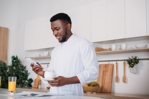 Hombre afroamericano con taza de café escribiendo en el teléfono inteligente en la cocina
 - Foto, Imagen