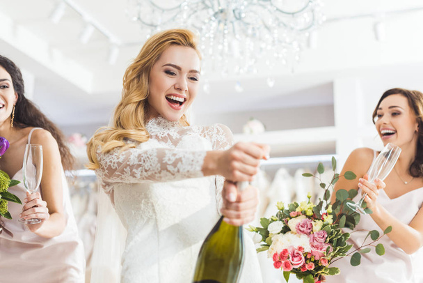 Молодые улыбающиеся невесты и подружки невесты открывают шампанское в магазине свадебной моды
 - Фото, изображение