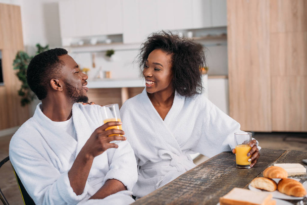 Afrikanisch-amerikanisches Paar hält Gläser mit Saft in der Hand und sitzt am Tisch mit Toasts und Croissants  - Foto, Bild