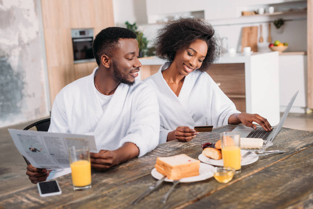 Young man with newspaper and smiling girlfriend with credit card doing online shopping - Photo, Image