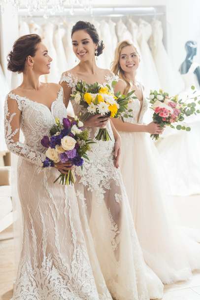 Smiling women in wedding dresses with flowers in wedding atelier - Фото, изображение