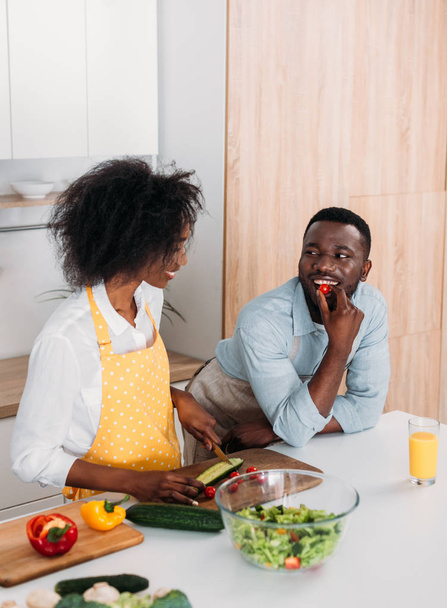 Young woman cutting cucumber on board and boyfriend eating cherry tomato - Fotó, kép