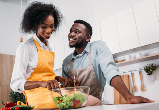 Low angle view of smiling woman mixing salad in bowl and sitting on table with boyfriend standing near - Φωτογραφία, εικόνα