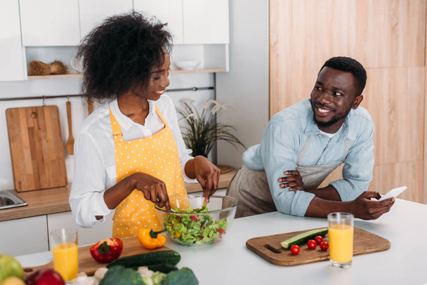 Smiling woman mixing salad in bowl at table and boyfriend standing with smartphone in hand - Fotó, kép