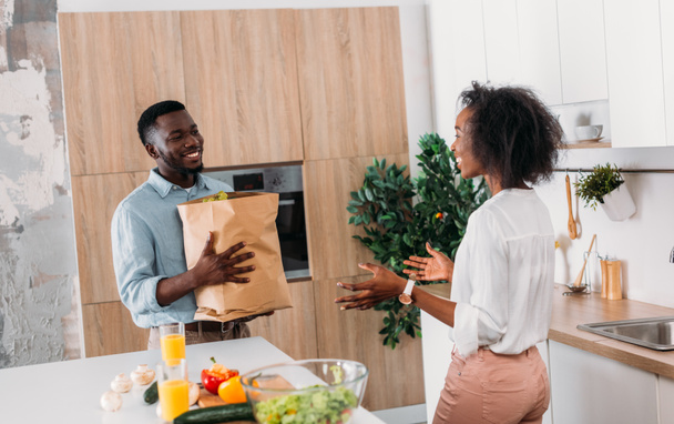 Jonge Afro-Amerikaanse man brengen van de papieren zak met producten terwijl vriendin permanent in keuken - Foto, afbeelding