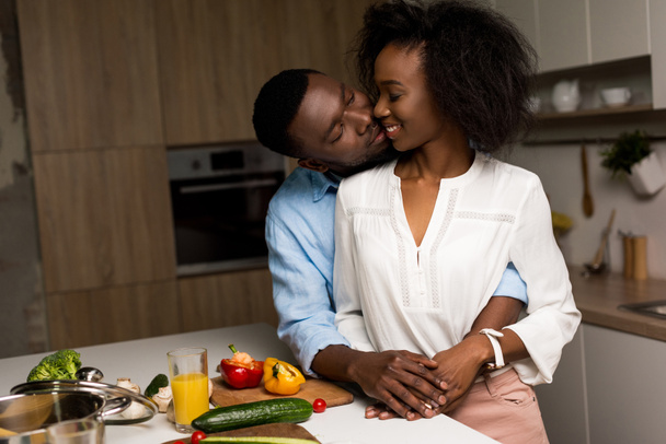 Hymyilevä afrikkalainen amerikkalainen pari halaa lähellä pöytää vihannesten ja kattilan kanssa
 - Valokuva, kuva