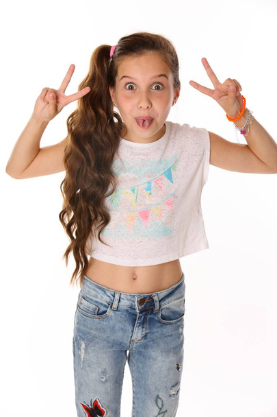 Portrait of happy slender cheerful teenage girl. The child elegantly poses makes funny faces and shows her tongue. Young fashionista in blue jeans and with a bare belly. - Photo, Image
