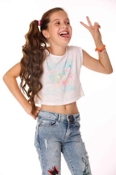 Portrait of happy slender cheerful teenage girl. The child elegantly poses makes funny faces and shows her tongue. Young fashionista in blue jeans and with a bare belly. - Photo, Image