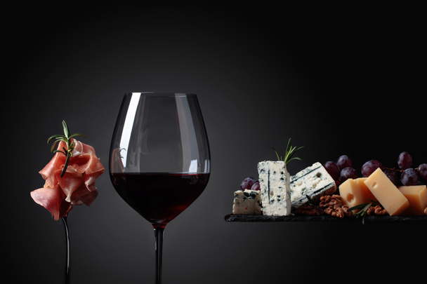 Glass of red wine with various cheeses , grapes and prosciutto . - Photo, Image