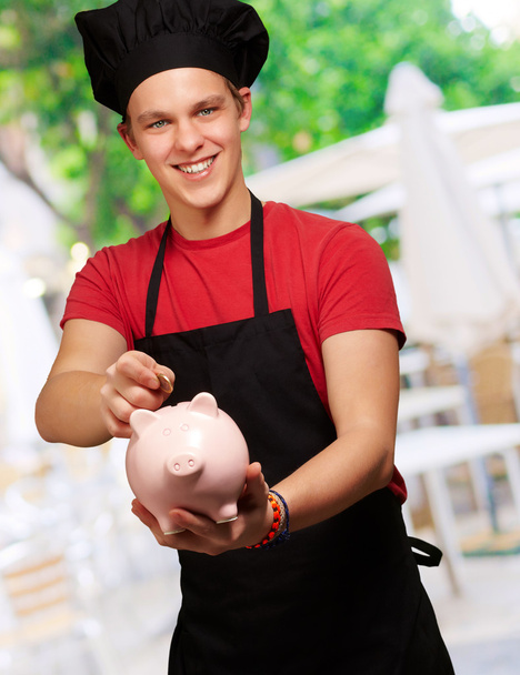 male chef holding a piggybank and a coin - Zdjęcie, obraz