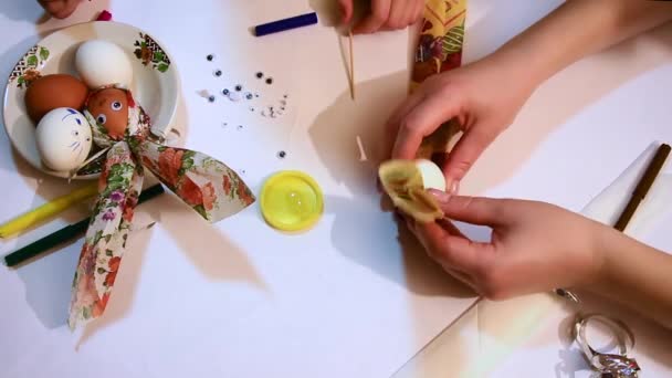 A close-up of a girls hands decorates a white egg with a colored napkin and a shiny ribbon. Funny faces. Decoration for Easter. - Materiał filmowy, wideo