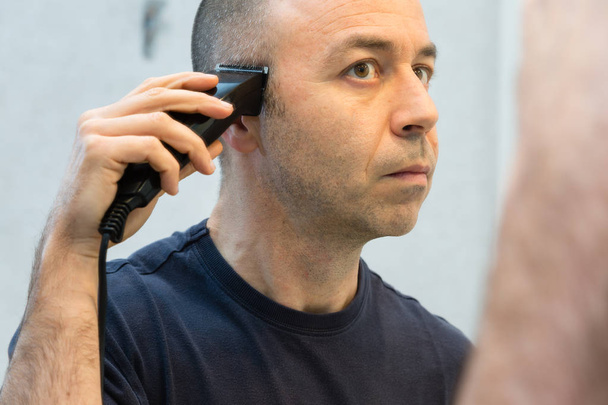 Kopasz férfi fekete póló borotválkozás a haját, elektromos borotva, a tükör előtt - Fotó, kép
