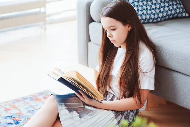 smart kid girl reading interesting book  at home, sitting with couch on background. Learning and education concept - Foto, Imagem