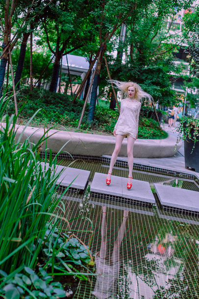Beautiful young blonde smiling happy woman with red lips in red shoes in a white dress play near a fountain in a summer day - Фото, зображення