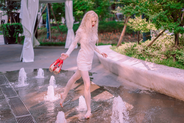 Mooie jonge blonde lachende gelukkige vrouw met rode lippen in rode schoenen in een witte jurk spelen in de buurt van een fontein in een zomerdag - Foto, afbeelding