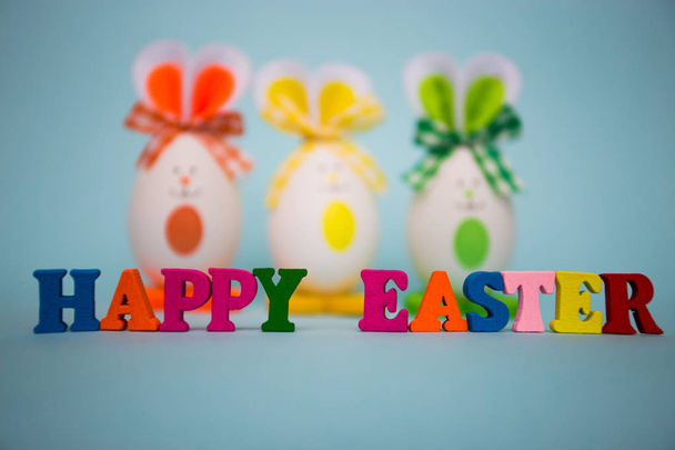 Feliz texto de Pascua de coloridas letras de madera con huevos divertidos en forma de lindo conejito sobre fondo azul. Feliz Pascua. Decoración festiva
 - Foto, Imagen