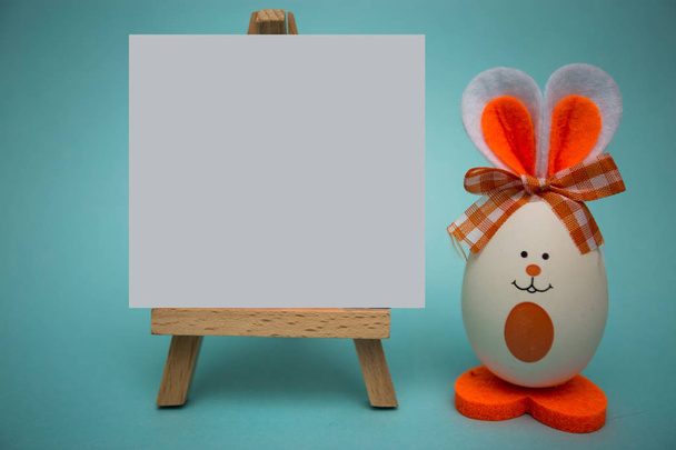 Papel blanco en blanco sobre caballete con huevo divertido en forma de lindo conejito sobre fondo azul. Feliz Pascua. Decoración festiva
 - Foto, Imagen