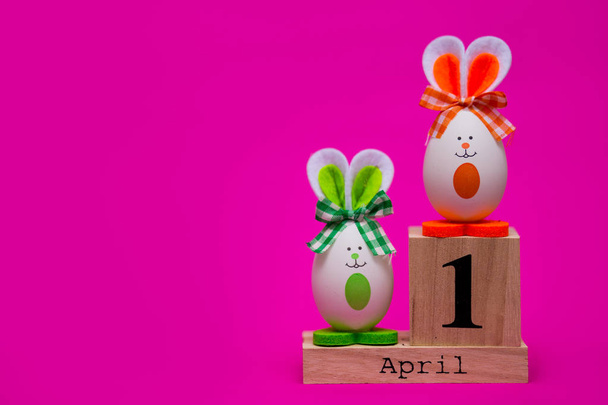 Разноцветные смешные яйца в виде милых кроликов с деревянным календарем, установленным на 1 апреля изолированы на розовом фоне. Счастливой пасхальной открытки. Апрель дурацкий день. Праздничное оформление
 - Фото, изображение