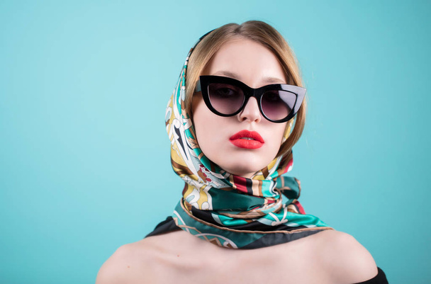 Close up shot of stylish young woman in sunglasses and you colored shawl, headscarf, scarf against blue background. Beautiful female model - Фото, изображение