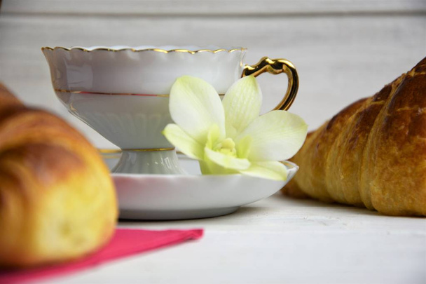 Uma xícara de café com café quente, croissants, guardanapo de mesa rosa e flor de orquídea verde em um fundo pintado de madeira
 - Foto, Imagem
