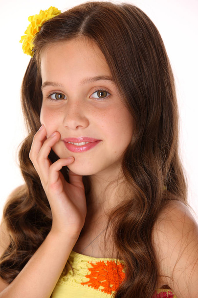 Close-up portrait of a beautiful charming happy young teenage girl in a yellow top and flower-pin. Pretty brunette child with chic long hair looks closely and smiles slightly. - Fotoğraf, Görsel