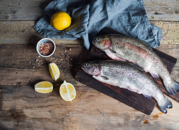 Two raw rainbow trout fish on a board on an old wooden background and lemon and pink sea salt in a small bowl. - Valokuva, kuva