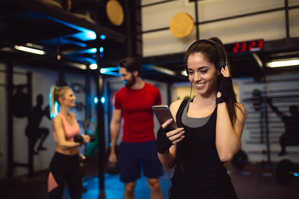 Sportswoman with smartphone listening to music in gym. Friends are talking behind, blurred in background. - Foto, afbeelding
