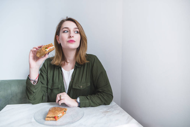 Όμορφη κοπέλα, κάθεται στο τραπέζι, τρώγοντας ένα σάντουιτς και αναζητούν μακριά. Γυναίκα με σάντουιτς φόντο γκρίζο τοίχο - Φωτογραφία, εικόνα
