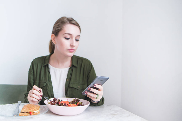 A pretty girl uses a smearphone while eating at the restaurant. A woman eats a salad and looks at a smartphone - Photo, image