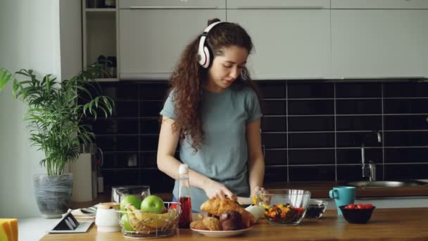 Attractive happy girl in headphones dancing and and listening to music while cooking healthy vegeatable salad in the kitchen at home at morning - Кадры, видео