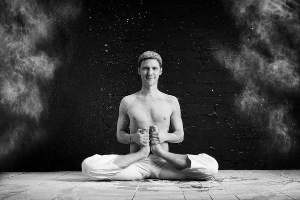 A man doing yoga in a white cloud of dust in a dark room. The concept of energy. Black-and-white photo. Hatha yoga. kandasana - Fotografie, Obrázek