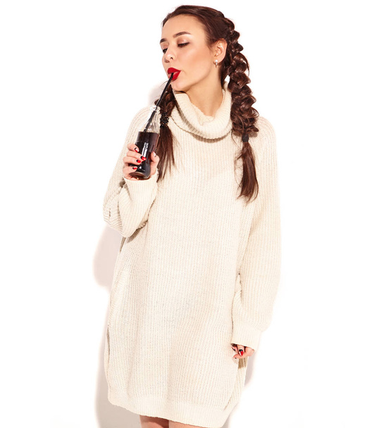 Young happy smiling woman model with bright makeup and red lips with two horns  in summer warm sweater clothes isolated on white. Drinking soda from glass bottle with straw - Fotoğraf, Görsel