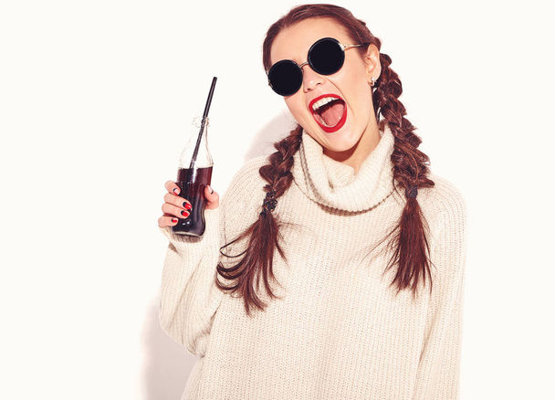 Young happy smiling woman model with bright makeup and red lips with two horns in summer warm sweater clothes and sunglasses isolated on white. Drinking soda from glass bottle with straw - Foto, immagini