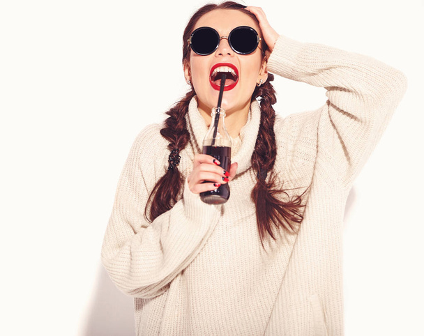 Young happy smiling woman model with bright makeup and red lips with two horns in summer warm sweater clothes and sunglasses isolated on white. Drinking soda from glass bottle with straw - Photo, Image