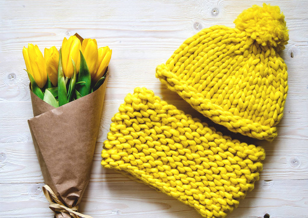 Um chapéu de malha amarelo e um ronco de fio Merino grosso. Nas proximidades é um buquê de tulipas amarelas
. - Foto, Imagem
