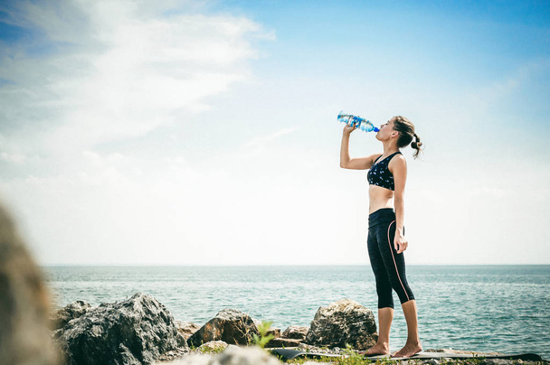 athletic woman drinking water from a bottle on the beach on a warm summer day, exercise in open air - Foto, afbeelding
