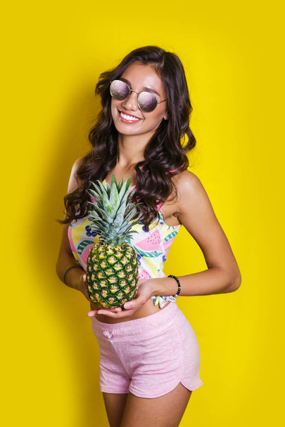 Fashion summer portrait smiling indian girl in sunglasses and pineapple over yellow background - Foto, immagini