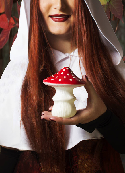 Pretty smiling redheaded girl holding an amanita mushroom on autumn nature background. - Photo, Image