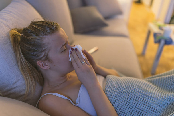 Αναστατωμένος νεαρή γυναίκα που υποφέρουν από γρίπη στο σπίτι και να φυσάει τη μύτη της κατά τη διάρκεια του πυρετού.  - Φωτογραφία, εικόνα