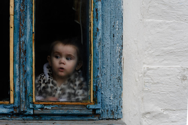 A little girl sits in an old hut on a window on which the paint faded - Φωτογραφία, εικόνα