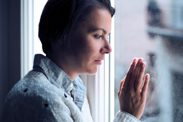 Vrouw naast een venster naar buiten op zoek in een eenzame bui - Foto, afbeelding