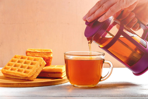 Female Hands Pour Tea from Brewer into Mug on White Wooden Table with Waffles. - Photo, Image